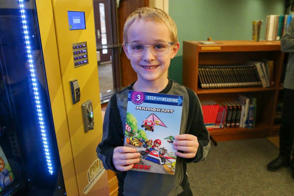 Young boy proudly holding a book in front of a vending machine.