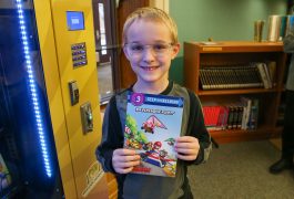 Young boy proudly holding a book in front of a vending machine.