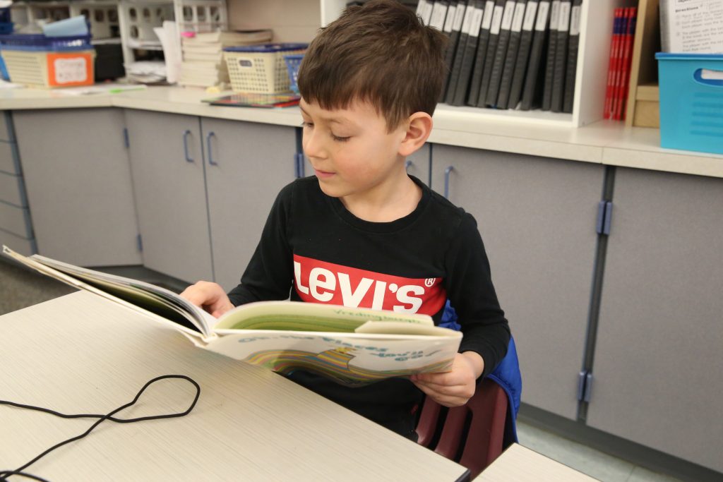 Young boy sitting at a table in a classroom, reading a book.