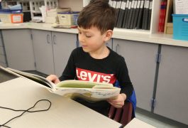 Young boy sitting at a table in a classroom, reading a book.