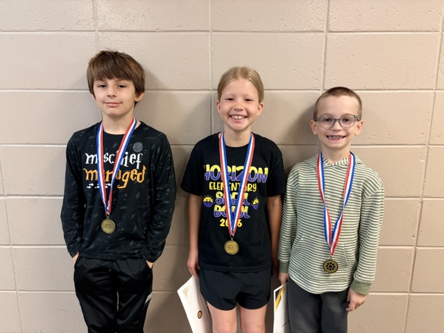 Three children posing in front of a wall with medals around their necks, each holding a medal, smiling and looking at the