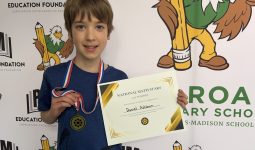 Young boy holding a medal in front of a sign for a school. The sign says "national mathematics".