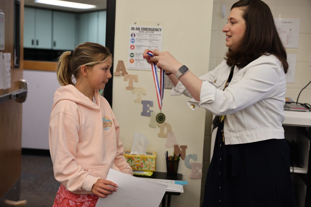 Woman in a white shirt pinning medal on girl's shirt.