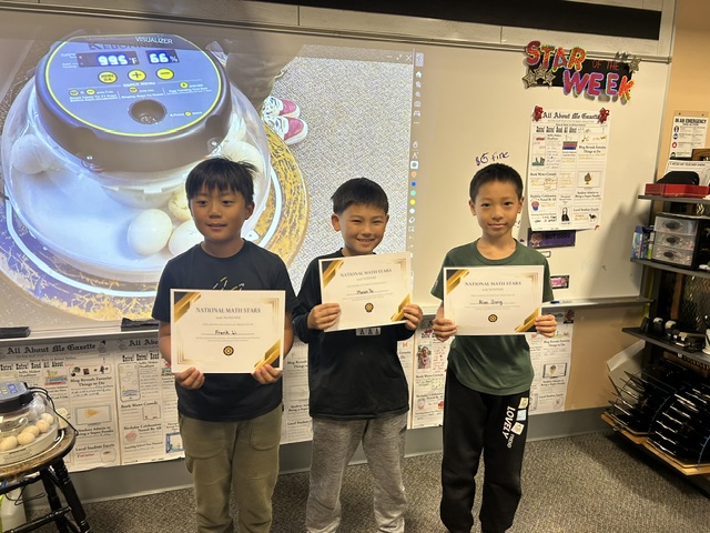 Three young boys proudly holding certificates in front of a classroom display.
