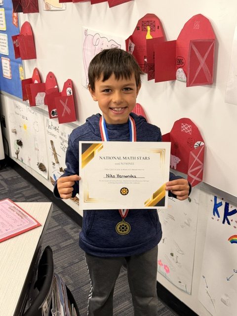 Young boy proudly holding a certificate of recognition in front of a bulletin board.