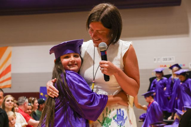 Two people hugging in a graduation gown ceremony, one person is speaking into a microphone.