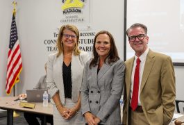 Three people standing in front of a podium at an event. The person on the left is wearing glasses and a red top. The middle