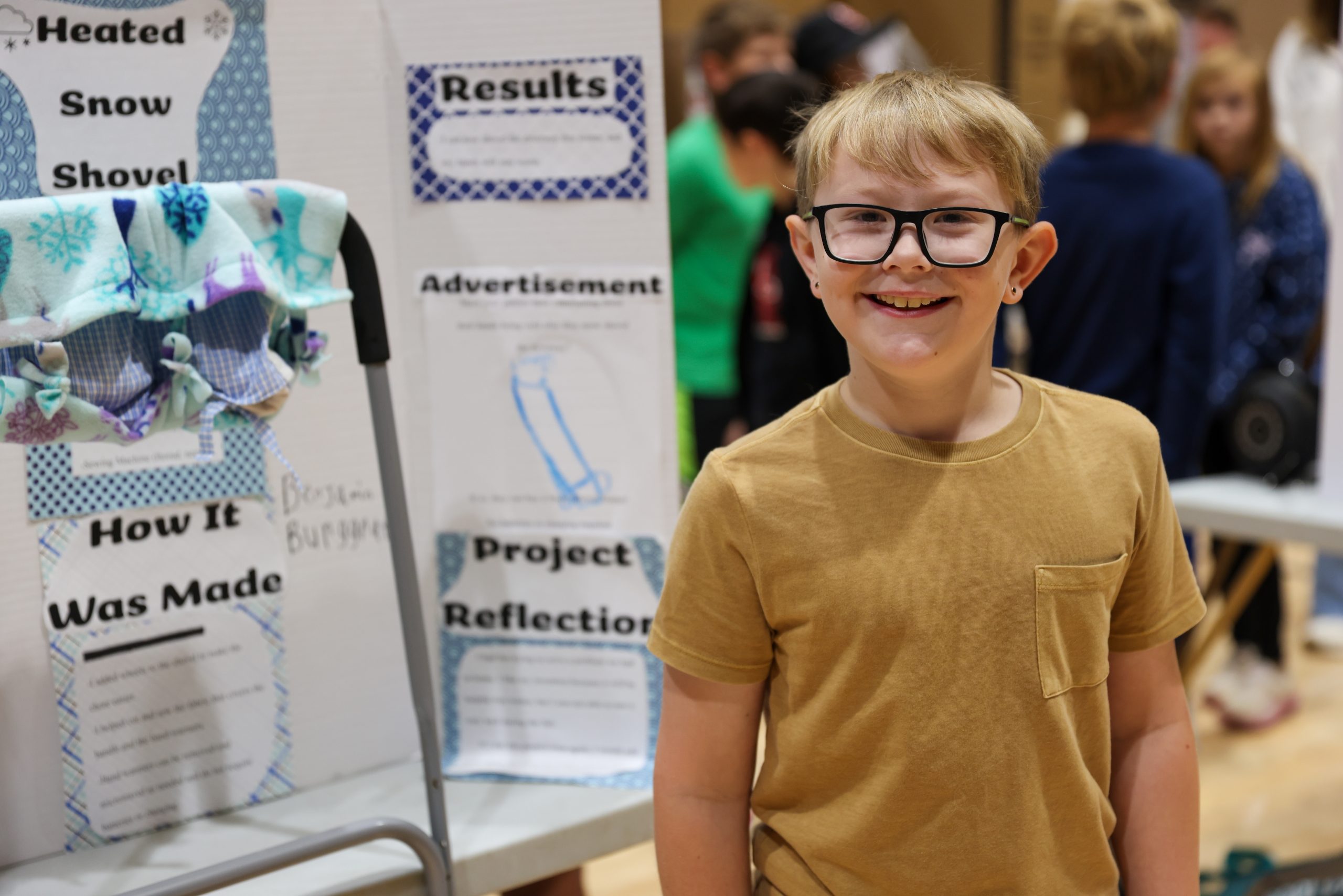 A young boy stands proudly in front of a table displaying his artwork, which includes a snowman and a reflection.